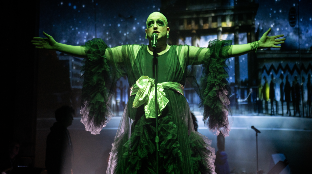 A performer dressed as Hedwig from "Hedwig and the Angry Inch" stands on stage with arms outstretched. They wear an elaborate green costume with ruffles and a large bow. Their face is dramatically made up with heavy eye makeup. The background shows a starry night sky, likely a digital projection. The lighting bathes the scene in a green glow, creating a theatrical and otherworldly atmosphere.